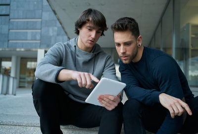 Front view of two young men talking sitting on a city staircase and lo