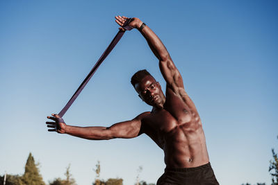 Male athlete doing resistance band exercise while standing against clear blue sky