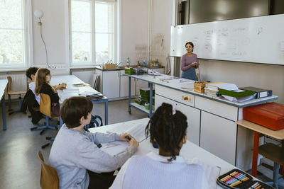 Smiling female teacher teaching students sitting at desk in classroom