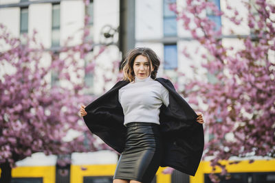 Portrait of beautiful woman holding winter coat against cherry trees in city