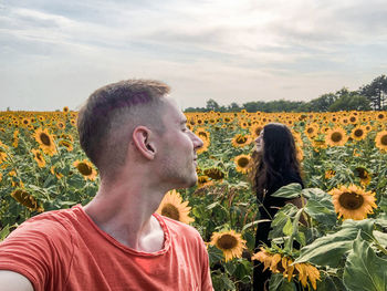 Portrait of young man and woman by sunflowers on field against sky