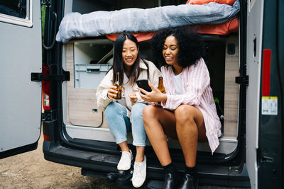 Multiracial young women with beer browsing on smartphone while sitting in van during road trip