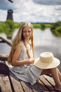 A teenage village girl in a dress stands at a wooden house by the river in summer