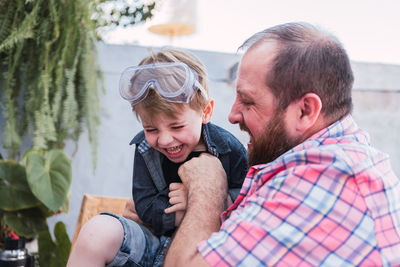 Unrecognizable bearded dad in checkered shirt playing with cheerful boy in safety glasses while sitting in daytime