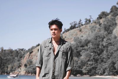 Young man looking away while standing at beach against mountain during sunny day