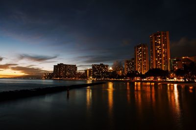 Illuminated buildings by river against sky at night