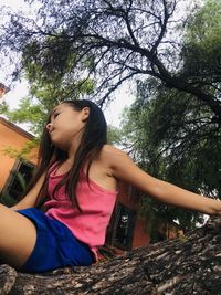 Beautiful young woman looking down while sitting on tree against plants