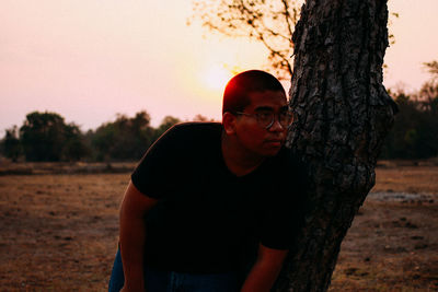 Portrait of young man standing by tree trunk against sky during sunset