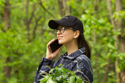 Smiling caucasian woman in black cap and glasses talking on a cell phone in the green spring forest.