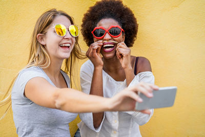 Smiling young woman taking selfie from mobile phone with friend against wall