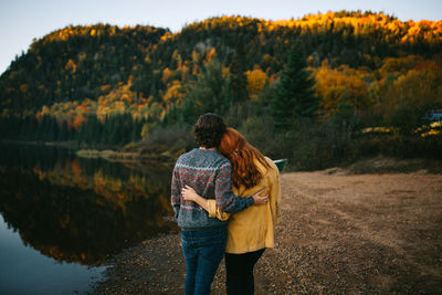 Back view of romantic unrecognizable traveling couple in stylish clothes embracing and admiring scenic autumn landscape of calm lake surrounded by lush forest during trip in canada