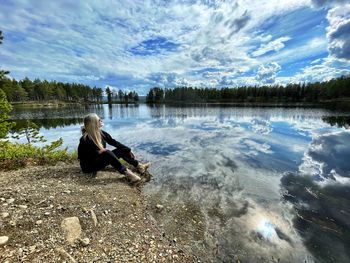 Side view of man sitting by lake against sky