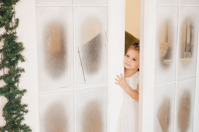 A little girl in a white dress peeps from behind the door. wide swing door decorated for christmas.