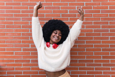 Young woman with arms raised standing against brick wall