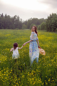 Mother and daughter in dresses and a hat stand in a field of yellow flowers in the summer day