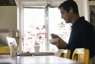 Side view of male freelancer having breakfast while doing video conference through laptop at home