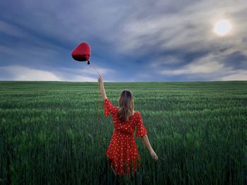 Woman in red dress standing in a field of green wheat and holding a red heart balloon