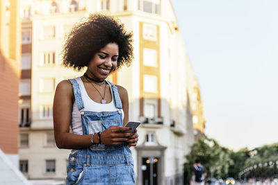 Smiling young woman using mobile phone against buildings