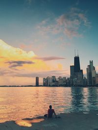 People on sea by city against sky during sunset