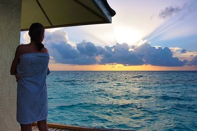 Rear view of woman wrapped in towel standing by sea during sunset