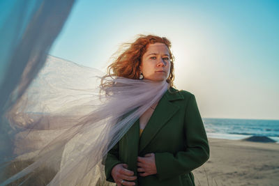Young woman looking away while standing on beach against sky