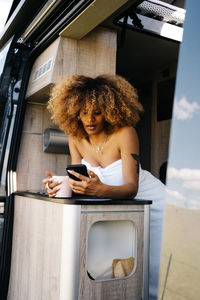Glad african american woman with mug of hot beverage smiling and browsing cellphone while resting in contemporary caravan in morning