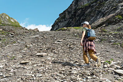 Rear view young woman in cap with backpack walking uphill along rocky trail in mountain hike, hiking