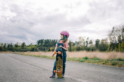 Young girl stood with a skateboard and helmet on a country road