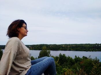 Side view of young woman sitting by lake against sky