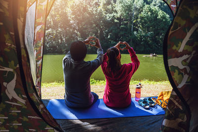 Rear view of people making heart shape with hands sitting on mat by lake