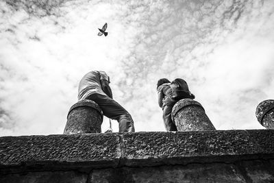 Low angle view of people sitting on bollard with bird flying against sky