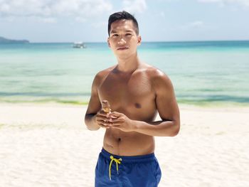 Portrait of shirtless man holding ice cream while standing at beach against sky