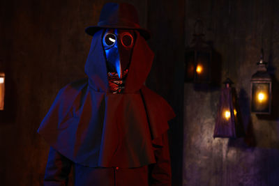 Portrait of a plague doctor in a black leather mask against a dark wall with lanterns.