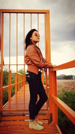 Side view of young woman standing on footbridge against sky