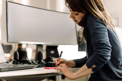 Female computer programmer talking on smart phone while writing at workplace