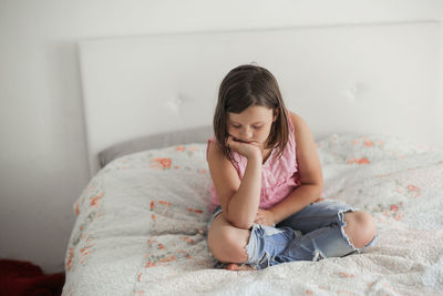 Girl child serious and frustrated sitting on a bed in a bright bedroom, concept age crisis 