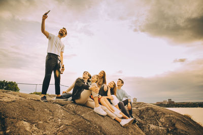 Man taking selfie with friends on rock formation against sky in picnic during sunset