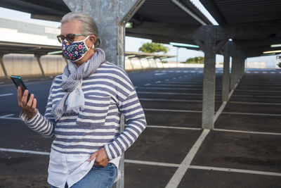 Senior woman wearing mask using phone standing in parking lot