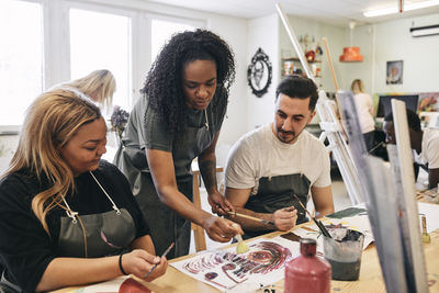 Mature female instructor teaching painting to young students at desk in art class