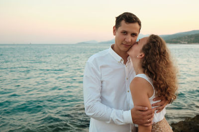 Romantic date on a sea coast. young adult woman kisses her beloved man against seaside sunset