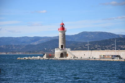 Lighthouse by sea against buildings and mountains