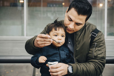 Father feeding baby food to son while sitting on bench in city