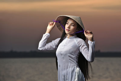 Beautiful young woman standing against sea at sunset
