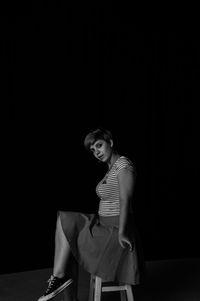 Portrait of young woman sitting on stool against black background