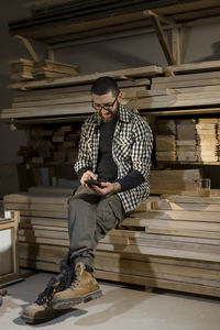 Carpenter using smart phone sitting on stacked planks at workshop