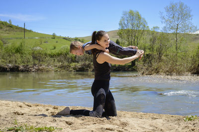 Full length of playful mother carrying son at riverbank