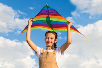 Girl holds a bright kite above her head against blue sky.