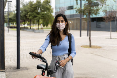 Beautiful woman wearing protective face mask while standing with bicycle in city during covid-19 pandemic