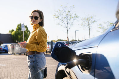 Smiling woman with smart phone standing by electric car at charging station