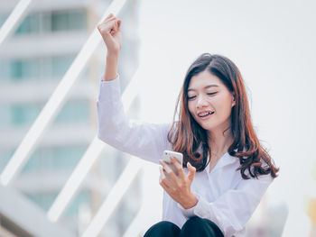 Low angle view of happy young woman using mobile phone while sitting outdoors